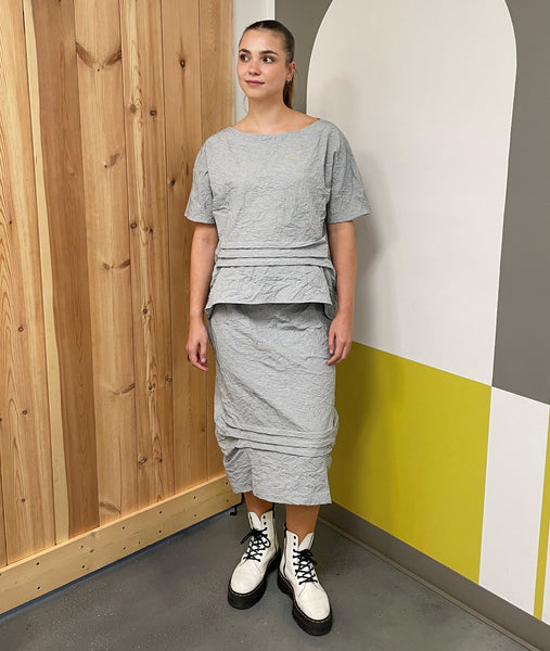 model in a grey and white pinstripe an elastic waist skirt with hip pockets and horizontal tucks at the bottom, creating a high-low hem, worn with a matching top with the same horizontal tucks at the waist
