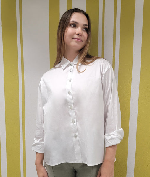 model in a green pant with a white button down blouse with a split at the sides and a lower hem in the back. model is wearing with rolled up sleeves