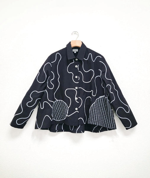 black cotton button down with a white swirl embroidered all over. blouse has long sleeves, a short full body, and constrasting check print circles in the front