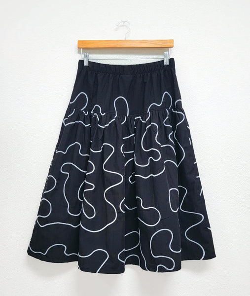 black elastic waist skirt with a white embroidery swirl design all over. skirt has gathered skirt attached to a slightly gathered panel at the hips