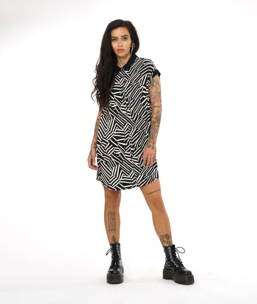 model in a short, cap sleeve button down shirt dress with a black collar and sleeve cuffs. Dress has a black and white broken stripe print and a curved hemline
