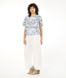 model in a straight leg white pant with a split at the ankle at the center front seam, worn with a boxy pullover top with a rounded neckline in a blue and black leaf print on a white background