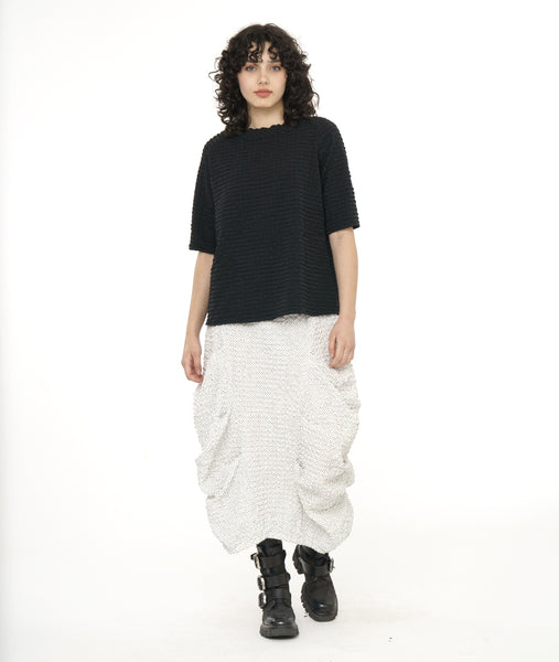model in a white and dot print skirt with large pleating along the sides, worn with a textured black pullover top with a short standing collar and 3/4 length raglan sleeves