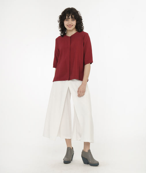 model in a wide leg white pant with an apron style panel overlay, worn with a brick red button up top with 3/4 sleeves and a round neckline
