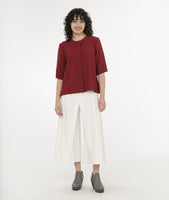 model in a wide leg white pant with an apron style panel overlay, worn with a brick red button up top with 3/4 sleeves and a round neckline