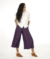 model in a wide leg purple pant with an apron style panel overlay, worn with a white button up top with 3/4 sleeves and a round neckline