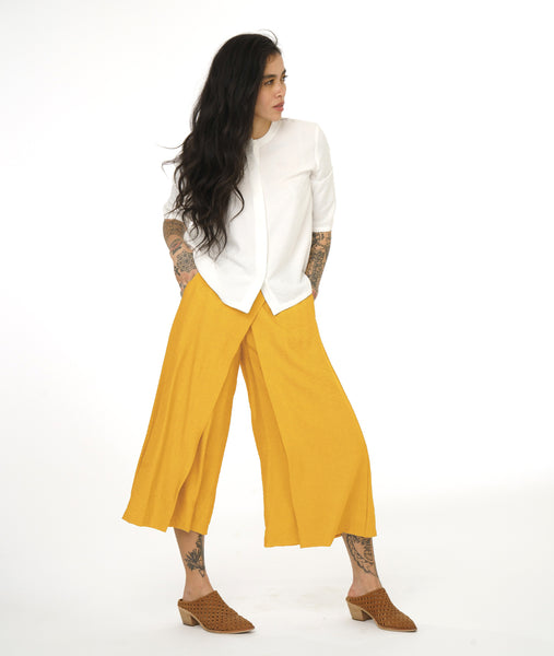 model in a wide leg saffron yellow pant with an apron style panel overlay, worn with a white button up top with 3/4 sleeves and a round neckline