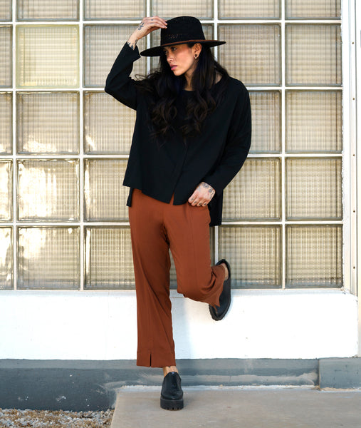 model in a slim rust color pant, worn with a boxy long sleeve black top with splits at the sides and center front. hem is longer in the back. worn outside with a black hat.