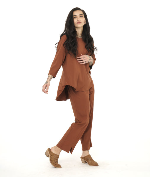 model in a slim rust color pant with a matching flowy pull over top. Top has 3/4 sleeves, a round neckline and a rounded hem with a high-low shape