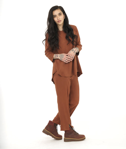 model in a slim rust color pant with a matching flowy pull over top. Top has 3/4 sleeves, a round neckline and a rounded hem with a high-low shape