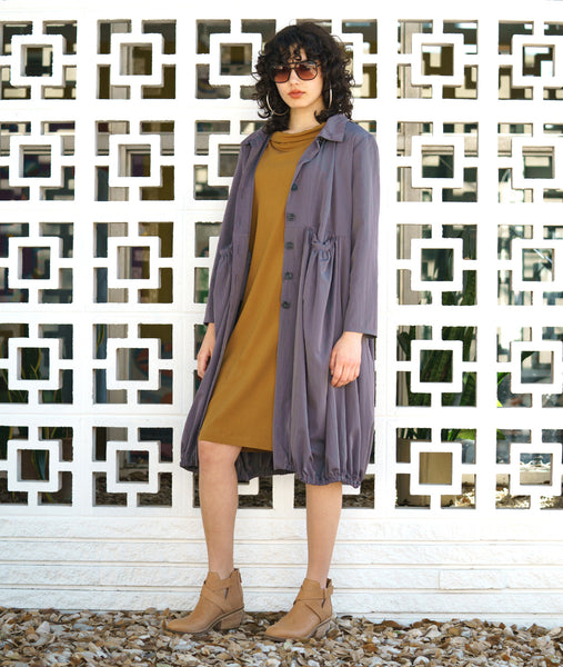 model in a ochre shift dress with a cowl neck, worn with a grey jacket with a gathered bottom hem and pockets, collar and long sleeves