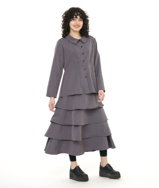model in a grey button down blouse with a pleated drape in the back, worn with black leggings and a long tiered skirt in a matching fabric