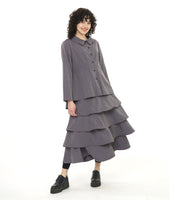 model in a grey button down blouse with a pleated drape in the back, worn with black leggings and a long tiered skirt in a matching fabric