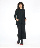 model in a black button down blouse with a standing collar, long sleeves and a round pocket at the hips. worn with a matching wide leg black pant.  fabric has a pinched circle texture