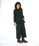 model in a black button down blouse with a standing collar, long sleeves and a round pocket at the hips. worn with a matching wide leg black pant.  fabric has a pinched circle texture