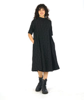 model in a black dress with 3/4 raglan sleeves, a short cowl neckline, and a flowy skirt hitting just below the knee. Dress has a textured circle design all over 