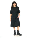 model in a black dress with 3/4 raglan sleeves, a short cowl neckline, and a flowy skirt hitting just below the knee. Dress has a textured circle design all over 