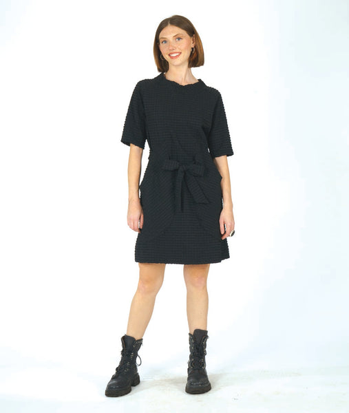 model in a textured black shift dress with a matching tie belt, deep exterior pockets, and elbow length raglan sleeves