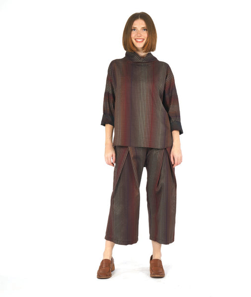 model in a pullover top with a dropped shoulder, rounded hem and oversized cowl neckline. worn with a matching pant with a double overlap at the top of each leg. top and pant are striped in varying shades of reds and browns