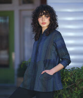 model in a button down blouse with long sleeves, a standing collar, and contrasting directional panels on the body. fabric is striped with varying shades of blues. worn with a slim leg black pant