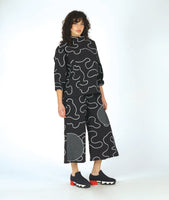 model in a wide leg black pant with contrasting round patches in a black and white grid print. worn with a matching pullover blouse with a standing cowl neckline, dropped shoulders, princess seams and a full boxy body. pants and top are black with a white swirl print embroidered all over