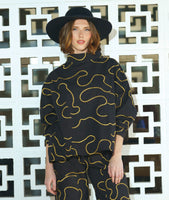 model in a wide leg black pant with cargo pockets, worn with a matching pullover blouse with a standing cowl neckline, dropped shoulders, princess seams and a full boxy body. pants and top are black with a yellow swirl print embroidered all over