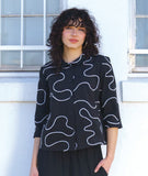 model in a slim black pant with a black button down blouse with 3/4 sleeves and a twin button detail. blouse has a white swirl print embroidered all over
