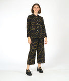 model in a wide leg pant with low pockets, worn with a matching blouse with long sleeves and a standing mandarin style collar. pants and blouse are both black with a yellow swirl pattern embroidered all over