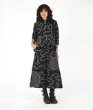 model in a black button down blouse with a low hem in back, 3/4 sleeves, circular black and white grid patches, and a white swirl print embroidered all over. worn with a matching wide leg pant with the same swirl pattern and patches
