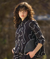 model in a black button down blouse with a low hem in back, 3/4 sleeves, circular black and white grid patches, and a white swirl print embroidered all over