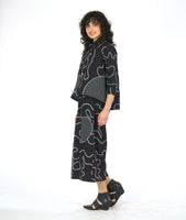 model in a black button down blouse with a low hem in back, 3/4 sleeves, circular black and white grid patches, and a white swirl print embroidered all over. worn with a matching wide leg pant with the same swirl pattern and patches