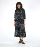model in a black button down blouse with a low hem in back, 3/4 sleeves, circular black and white grid patches, and a white swirl print embroidered all over. worn with a matching wide leg pant with the same swirl pattern and patches