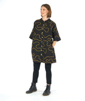 model in a black leggin with a black cocoon shape shirt dress with a yellow swirl print embroidered all over. dress has solid black cuffs and collar