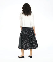 model in a white boxy button down blouse with a black skirt. skirt is gathered at the hip and is black with a white swirl print embroidered all over, and large circular patches with a black and white grid print