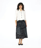 model in a white boxy button down blouse with a black skirt. skirt is gathered at the hip and is black with a white swirl print embroidered all over, and large circular patches with a black and white grid print