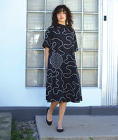 model in a bell shaped dress with pockets, 3/4 raglan sleeves, and a standing round collar. dress is black with a white swirl print embroidered all over, with round patches on black and white checker print