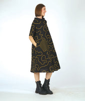 model in a bell shaped dress with pockets, 3/4 raglan sleeves, and a standing round collar. dress is black with a yellow swirl print embroidered all over, with round patches on black and yellow checker print