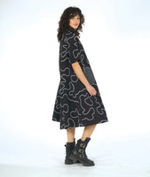 model in a bell shaped dress with pockets, 3/4 raglan sleeves, and a standing round collar. dress is black with a white swirl print embroidered all over, with round patches on black and white checker print