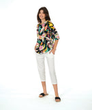 A model in a pullover shirt with a vibrant multicolor tropical floral print on a black background, paired with white cropped pants and black slide sandals, against a solid white studio background.