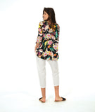 A model in a pullover shirt with a vibrant multicolor tropical floral print on a black background, paired with white cropped pants and black slide sandals, against a solid white studio background.