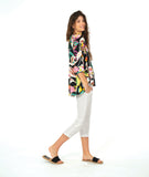 A model in a pullover shirt with a vibrant multicolor tropical floral print on a black background, paired with white cropped pants and black slide sandals, against a solid white studio background.