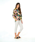 A model in a pullover shirt with a vibrant multicolor tropical floral print on a black background, paired with white cropped pants and black slide sandals, against a solid white studio background.