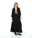 A woman modeling a black three-piece outfit consisting of a draped open-front jacket, wide-leg trousers, and a matching top, styled over a colorful floral print camisole and black block-heel sandals, against a plain white studio background.