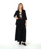 A woman modeling a black three-piece outfit consisting of a draped open-front jacket, wide-leg trousers, and a matching top, styled over a colorful floral print camisole and black block-heel sandals, against a plain white studio background.