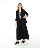 A woman modeling a black three-piece outfit consisting of a draped open-front jacket, wide-leg trousers, and a matching top, styled over a colorful floral print camisole and black block-heel sandals, against a plain white studio background.