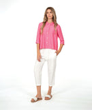 A woman modeling a bright pink button-up shirt with three-quarter sleeves, paired with white cropped pants and brown slide sandals, against a seamless white studio background.
