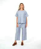 A woman modeling a light blue and white striped two-piece linen set consisting of a short-sleeve pullover top and matching cropped cargo pants, paired with brown loafers, against a plain white studio background.