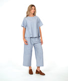 A woman modeling a light blue and white striped two-piece linen set consisting of a short-sleeve pullover top and matching cropped cargo pants, paired with brown loafers, against a plain white studio background.