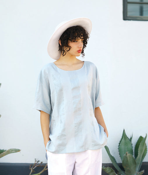 model in a straight leg white pant, white hat and a silvery blue pullover top with a large rounded neckline, elbow length raglan sleeve, and a large checkered print woven into the fabric