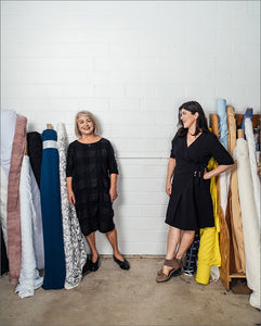 two women in black dresses standing in front of a white brick wall surrounded by tall rolls of fabric